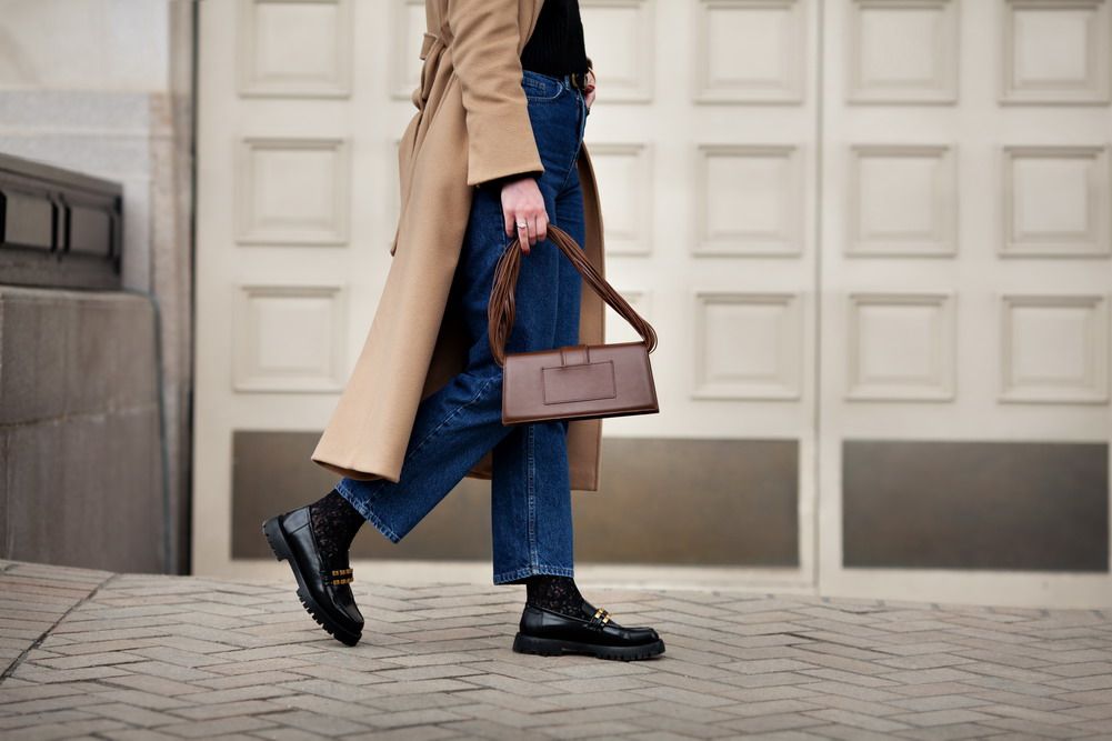 oman walking street in fashionable spring or autumn clothes cashmere coat, jeans, black loafers shoes and handbag. Female model in motion, street style fashion, close up legs